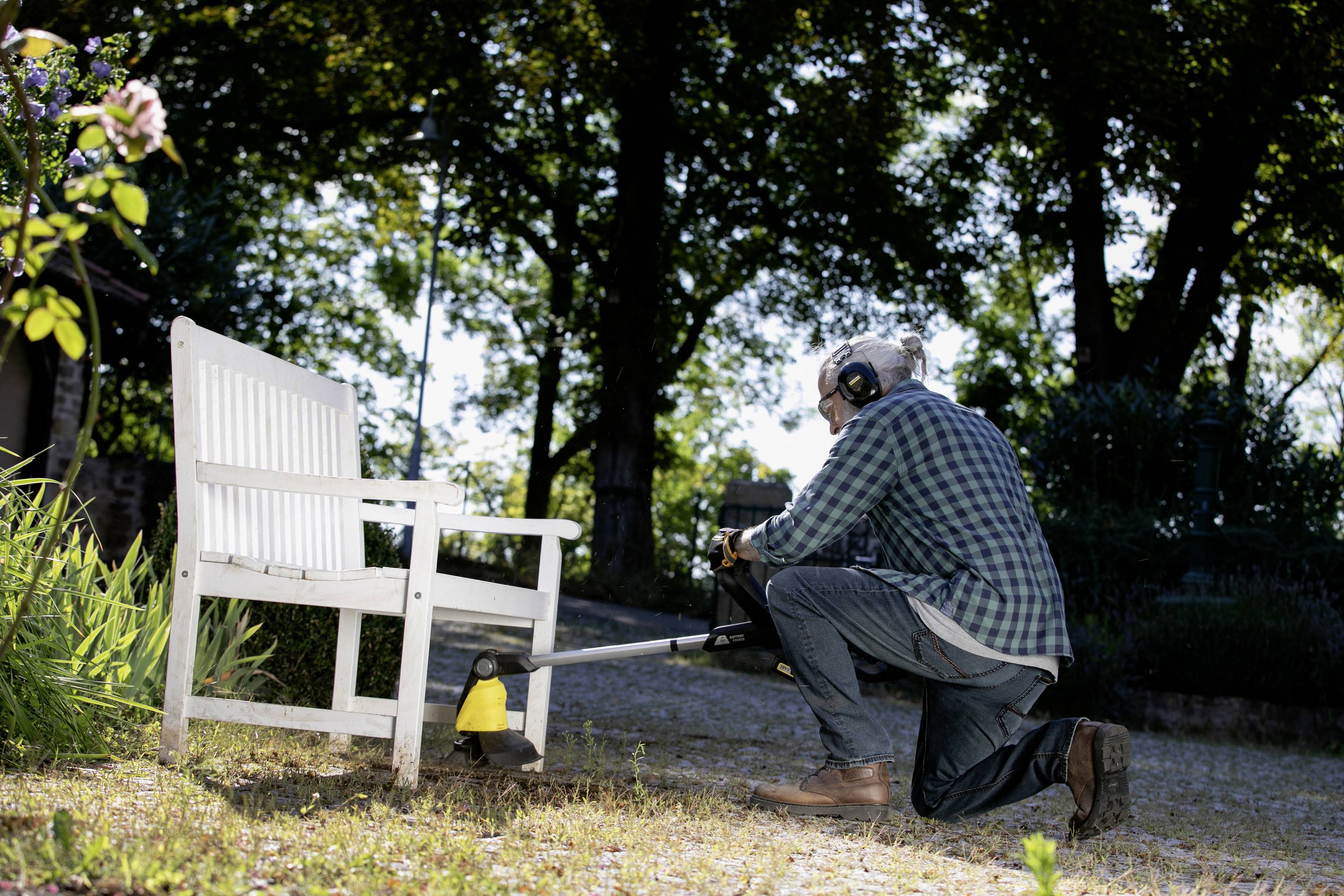 A man kneels on a gravel path and trims grass with a lawn strimmer next to a white garden bench in a shaded garden.