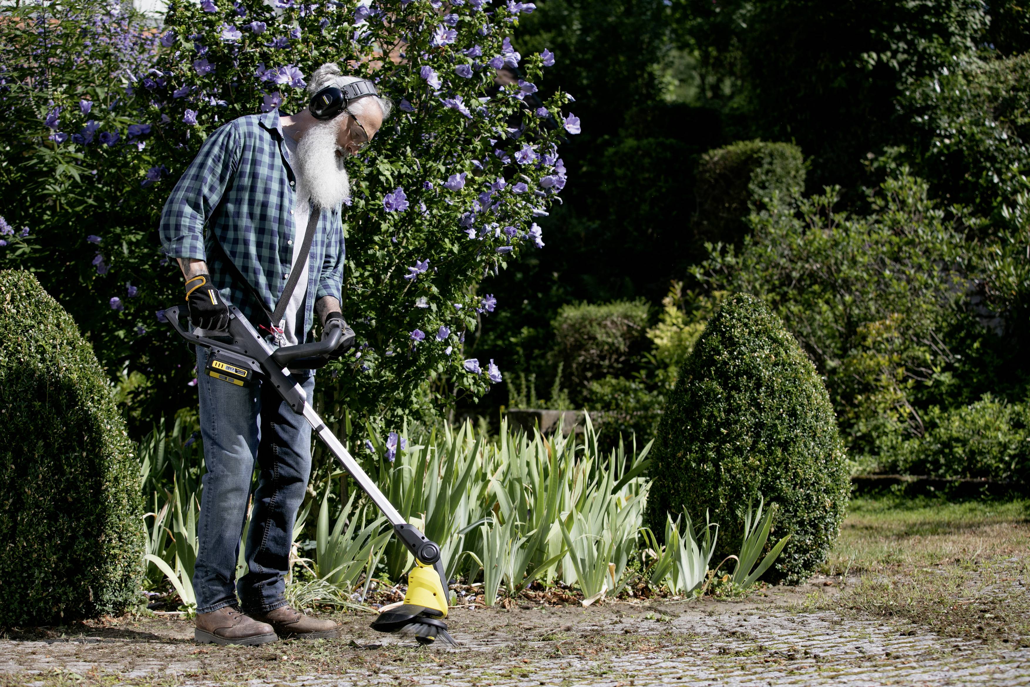 A person is using a lawn strimmer to clear pathways, surrounded by flowers and shrubs. They are wearing ear defenders.