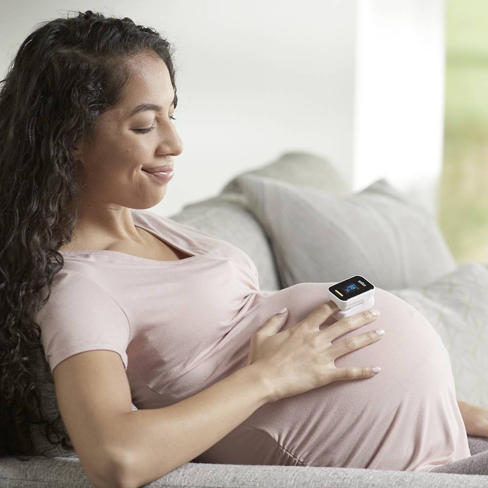 A pregnant woman sits smiling on a sofa, using a small pulse oximeter to monitor her blood and heart rate.