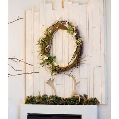 A rustic wreath of branches and flowers hangs on a white wooden wall above a mantelpiece decorated with pine branches.
