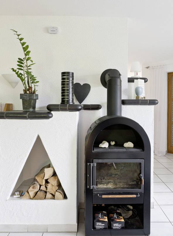 A modern wood-burning stove against a white wall. Wood is stacked in a compartment. Plants and decorations are placed on the mantelpiece.