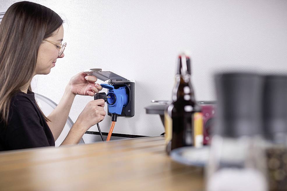 A woman is plugging an orange cable into a blue socket on the wall. Blurry objects are visible on a table in the foreground.