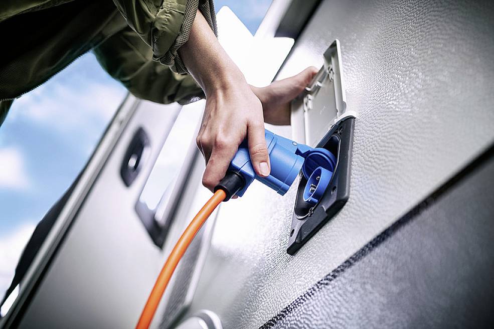 A person is connecting an orange charging cable to an electric vehicle. The focus is on the plug and the charging socket.