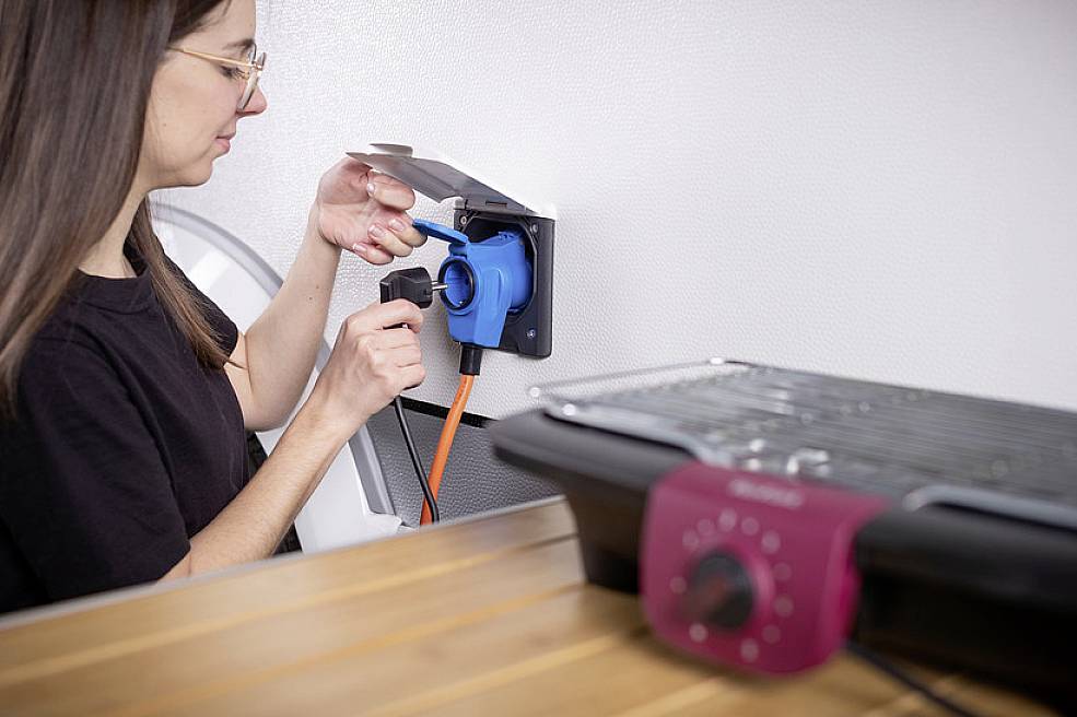 A person is plugging an electrical appliance into a blue socket, with a barbecue grill sitting on a wooden table in the foreground.