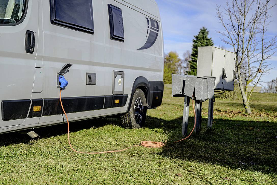 A motorhome is connected to an outdoor power supply via an orange cable. It is parked on a meadow.