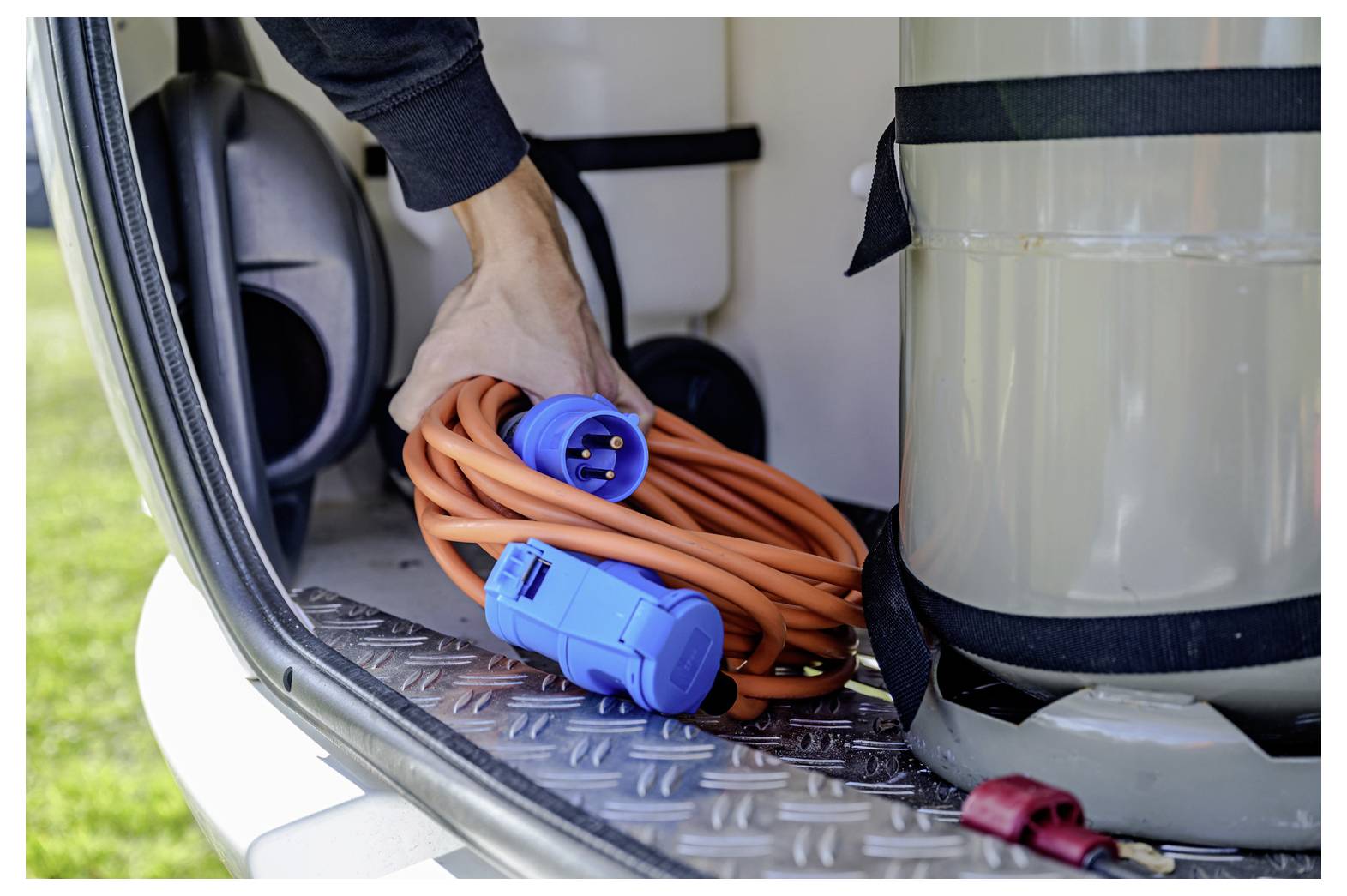 A person's hand is seen organizing an orange power cable with blue connectors inside a vehicle's trunk, next to a large silver tank.