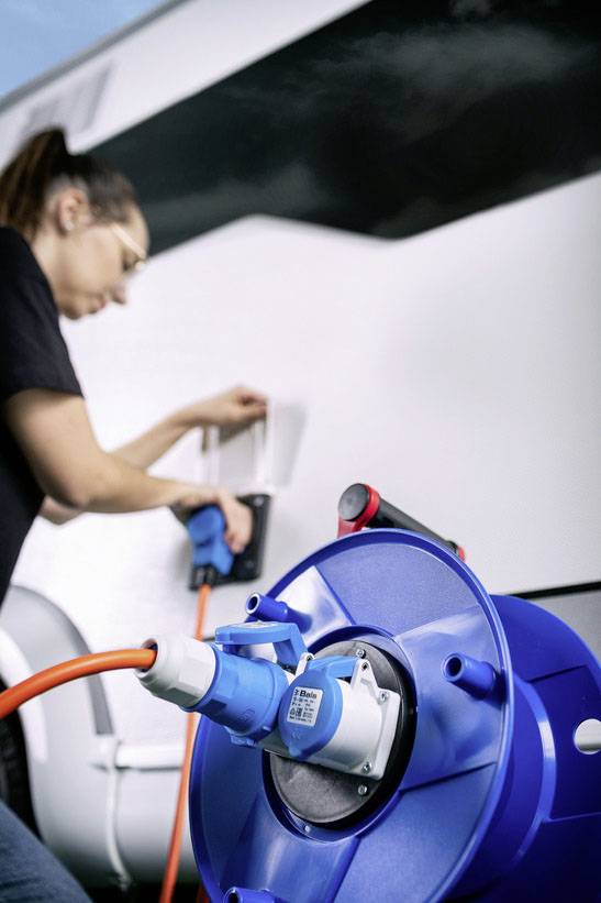 A person is plugging an electrical cable into a motorhome, which is connected to a blue power reel.