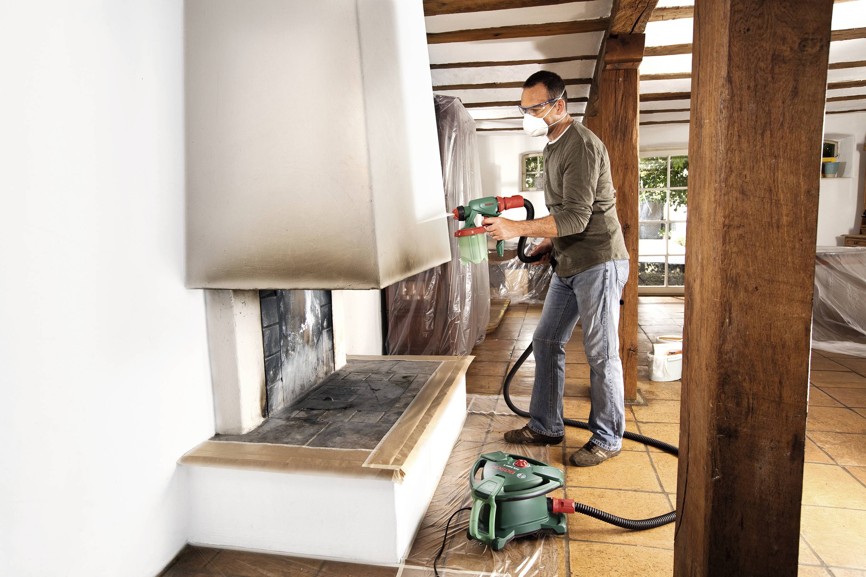 A man is using a spray gun to paint a white wall in a living room. The room is covered with plastic sheeting.
