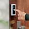 A hand presses the buzzer button of a modern, rectangular door intercom on a wooden door.