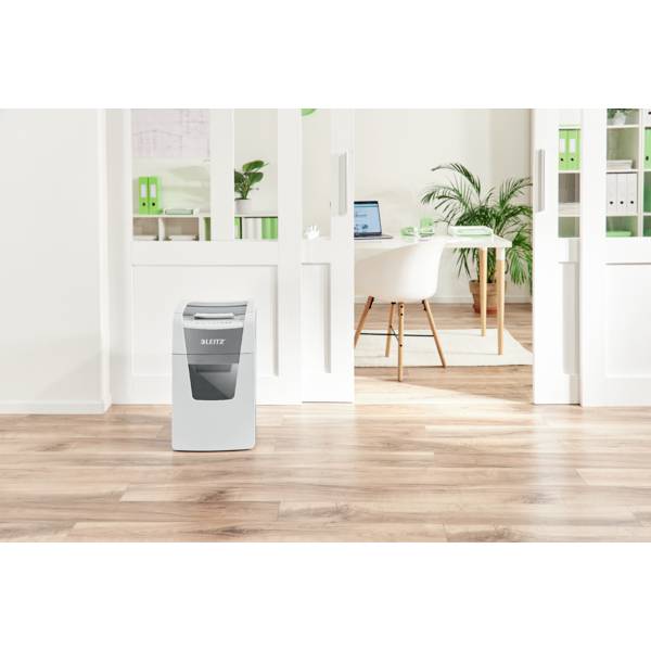 A shredder stands in a modern office with a wooden floor. In the background, a desk with a laptop and plants can be seen.