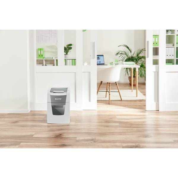 A paper shredder stands on a wooden floor in front of an open sliding door. In the background, an office can be seen with plants and a laptop.