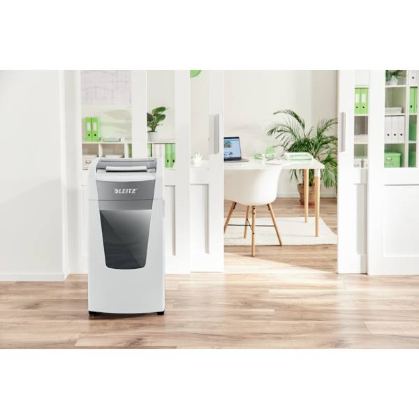 A mobile air purifier stands in a modern office with plants, large windows, and white furniture. A desk with a laptop is visible in the background.