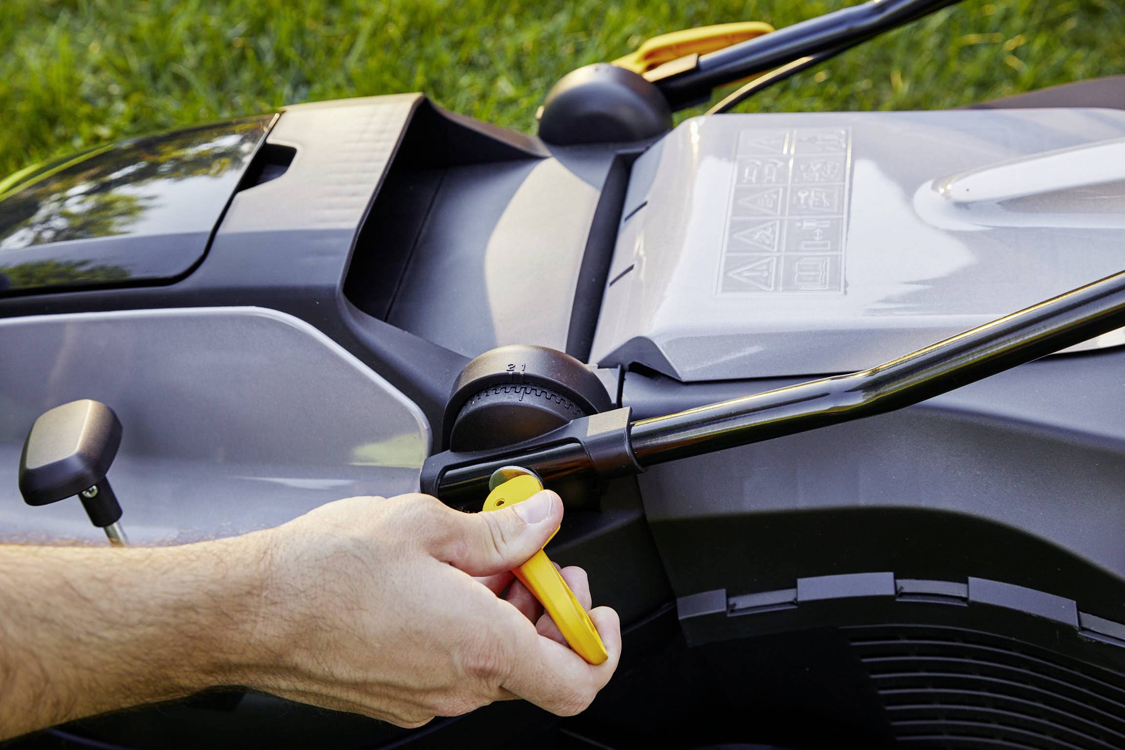A hand turns the starter of a lawnmower. The lawnmower is standing on green grass and shows details of the machine's construction.