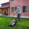 A man is mowing the lawn in front of a house with an electric lawnmower. In the background, a woman is sitting on the veranda.