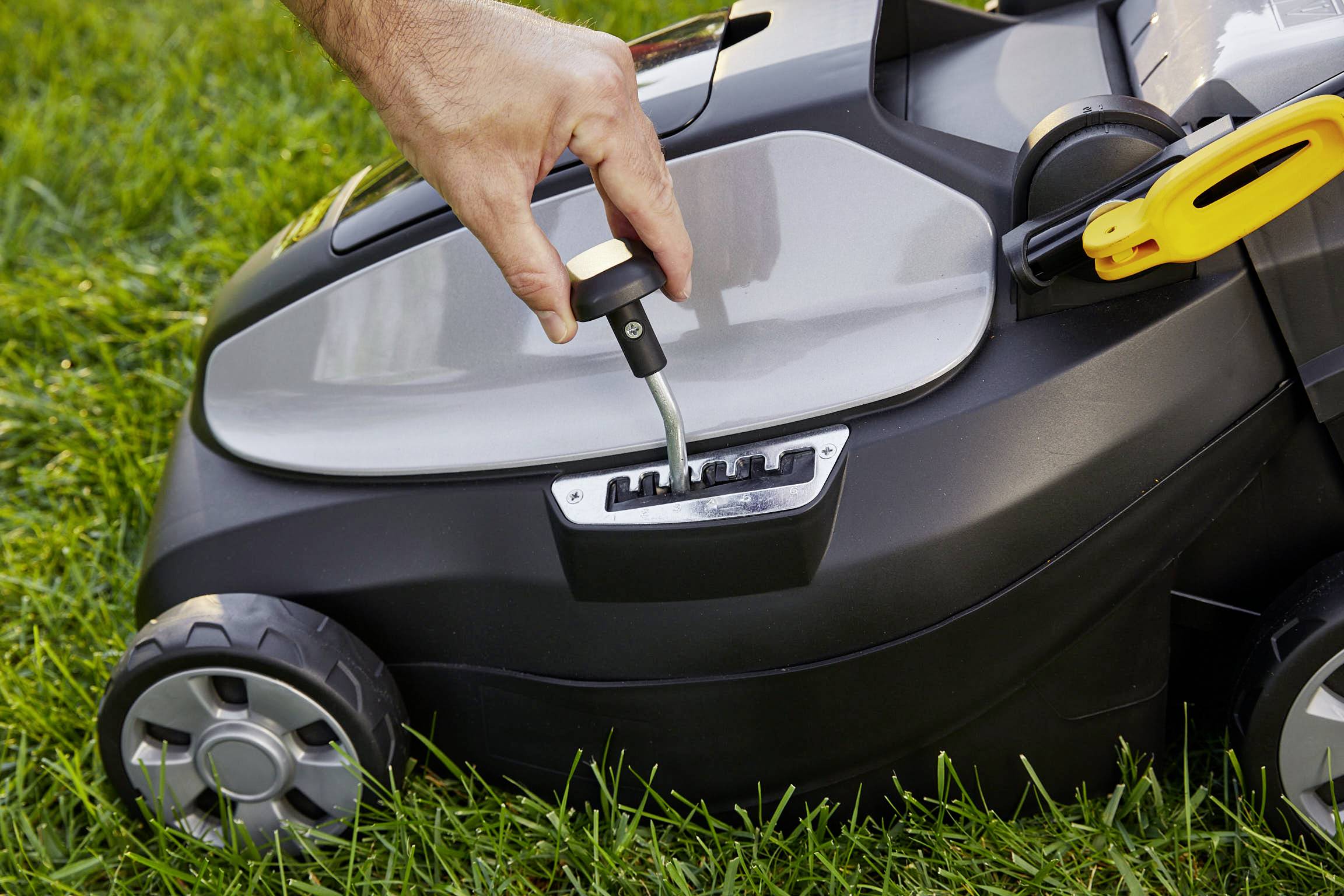 A lawnmower on a lawn. A hand adjusts the cutting height using a lever on the machine.