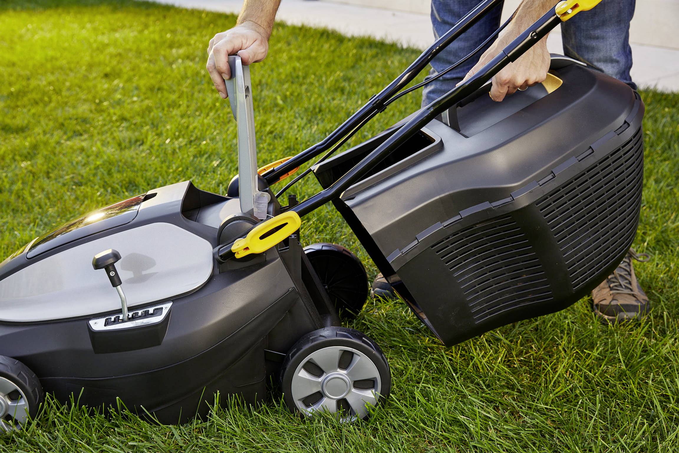 A person is securing the grass collection basket to a lawnmower on a green lawn.