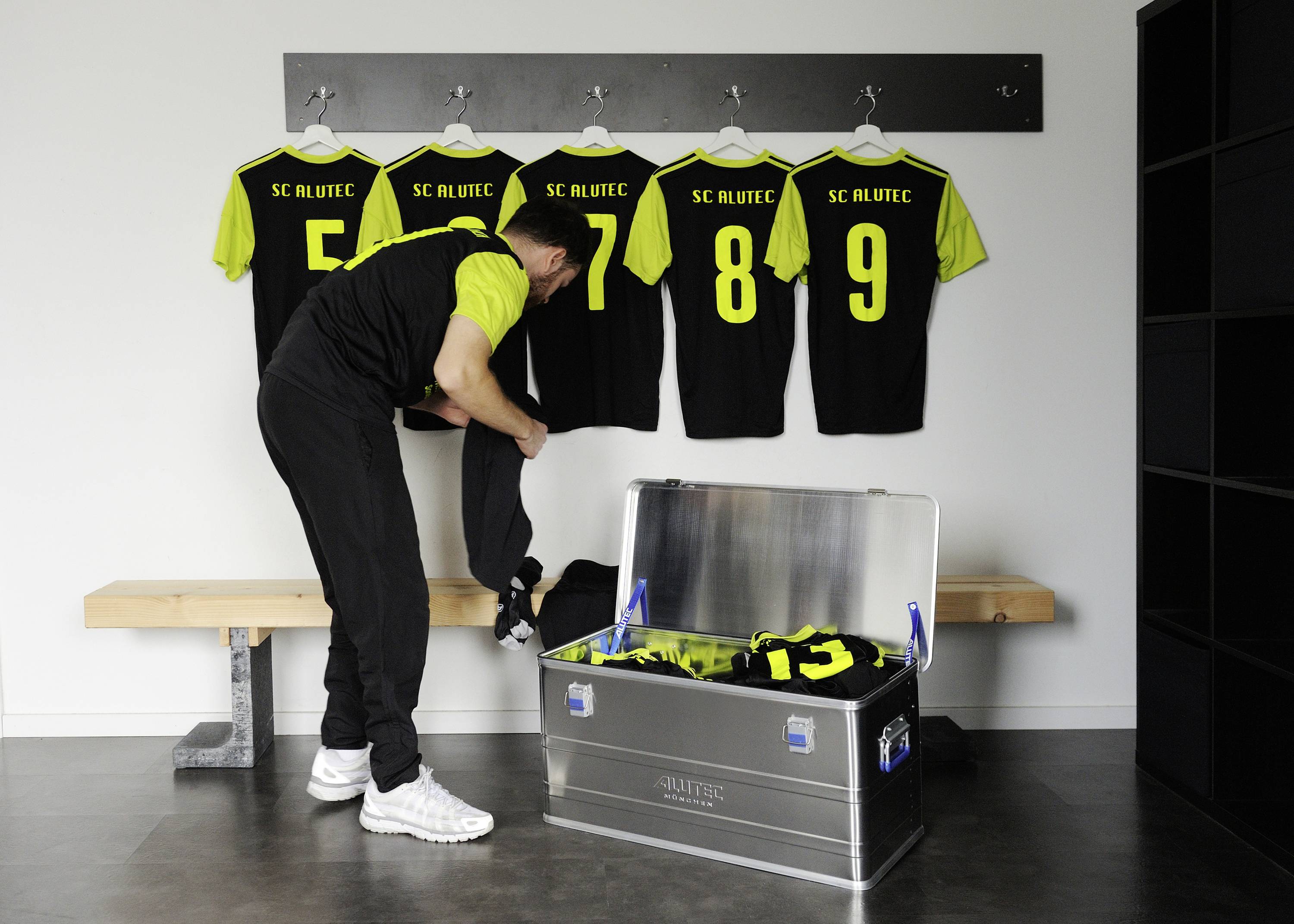 A man in the changing room is unpacking sports clothing from a metal locker. Four jerseys with yellow numbers are hanging on the wall.