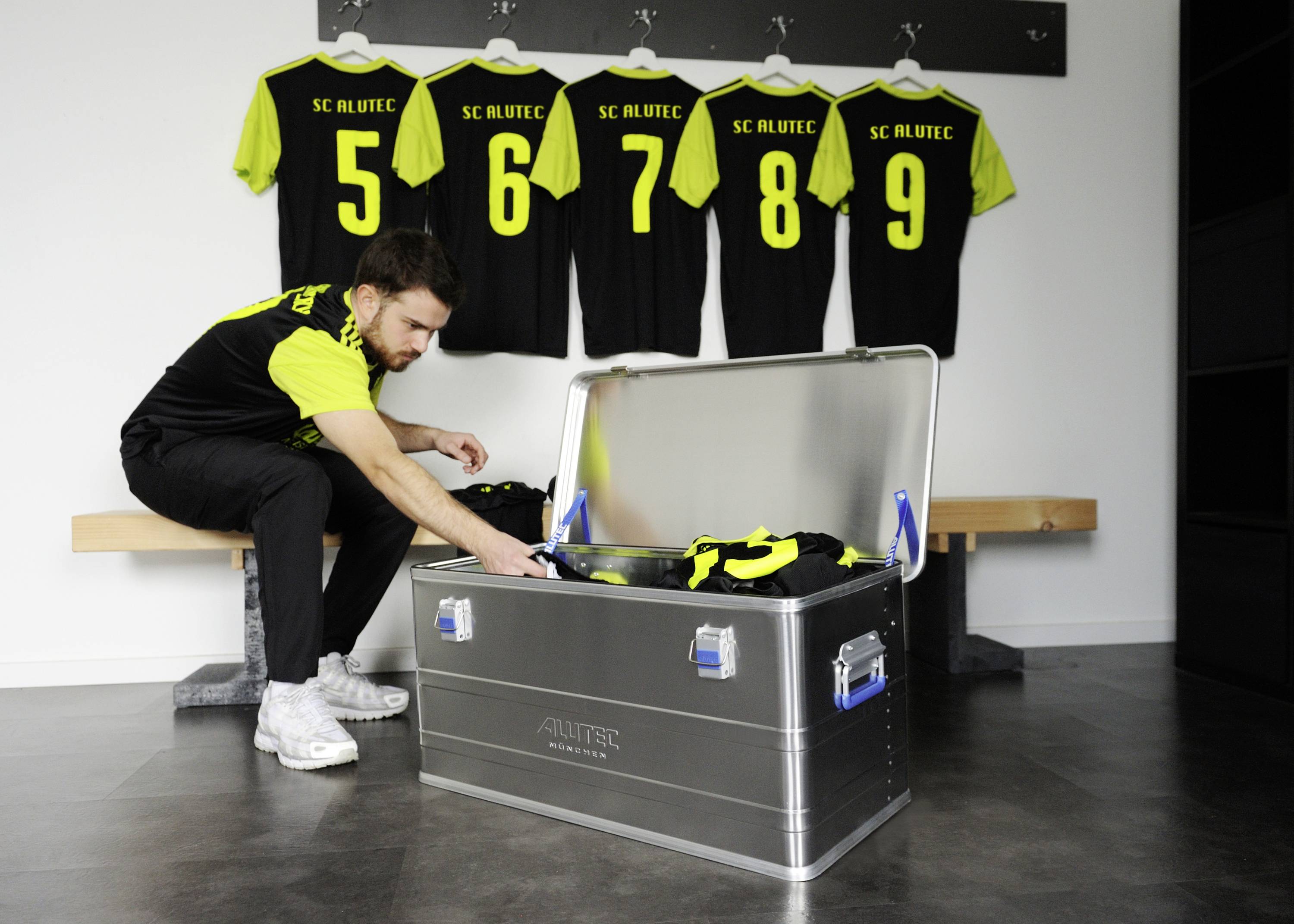 A man is unpacking jerseys from a metal box onto a wooden bench. In the background, four jerseys with numbers are hanging up.