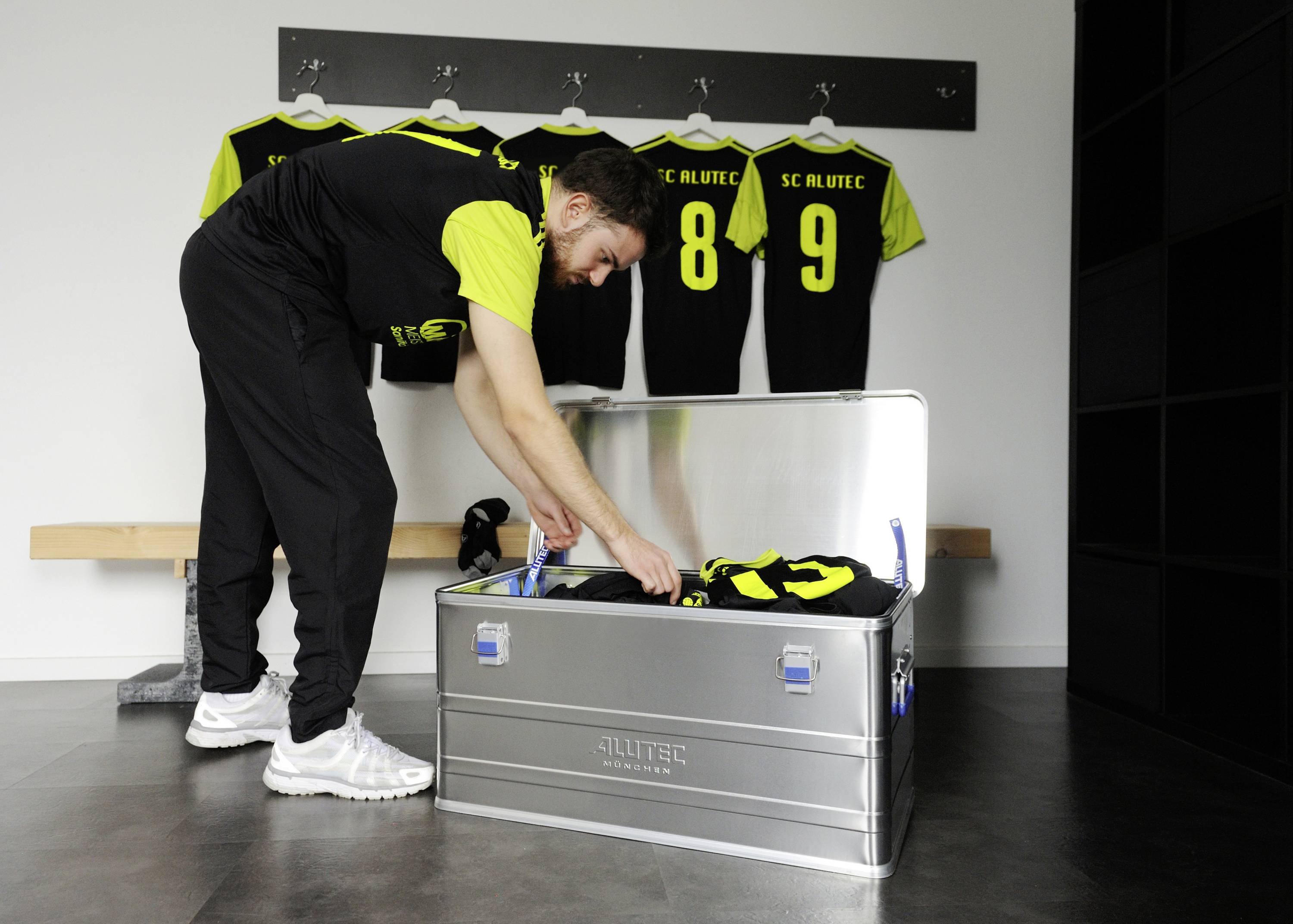 A man is sorting clothes in a metal box in a changing room. Sports jerseys with numbers are hanging on hooks in the background.