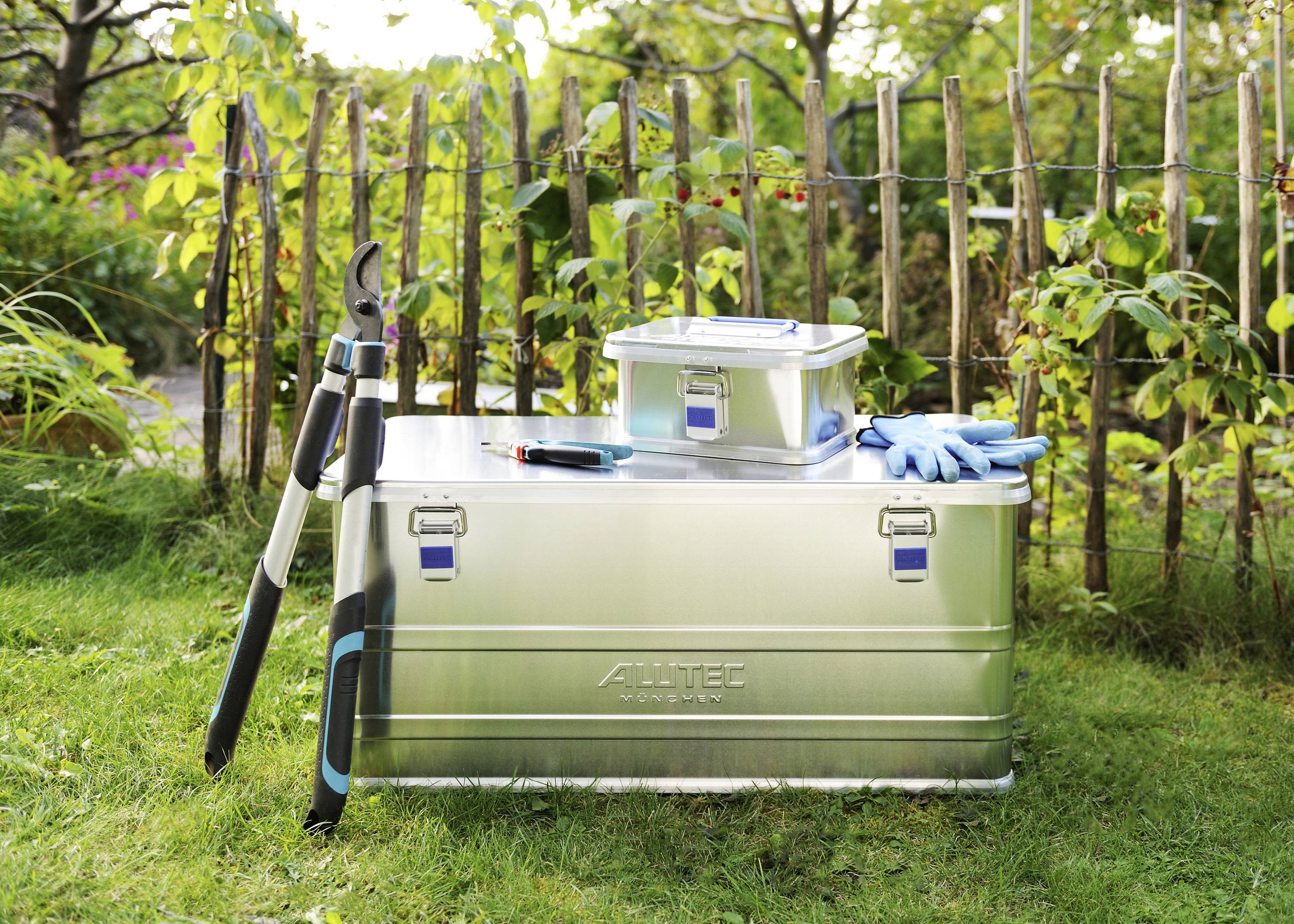 Garden tools and storage boxes on a lawn in front of a wooden fence. Contains secateurs, gloves, and a box, surrounded by plants.