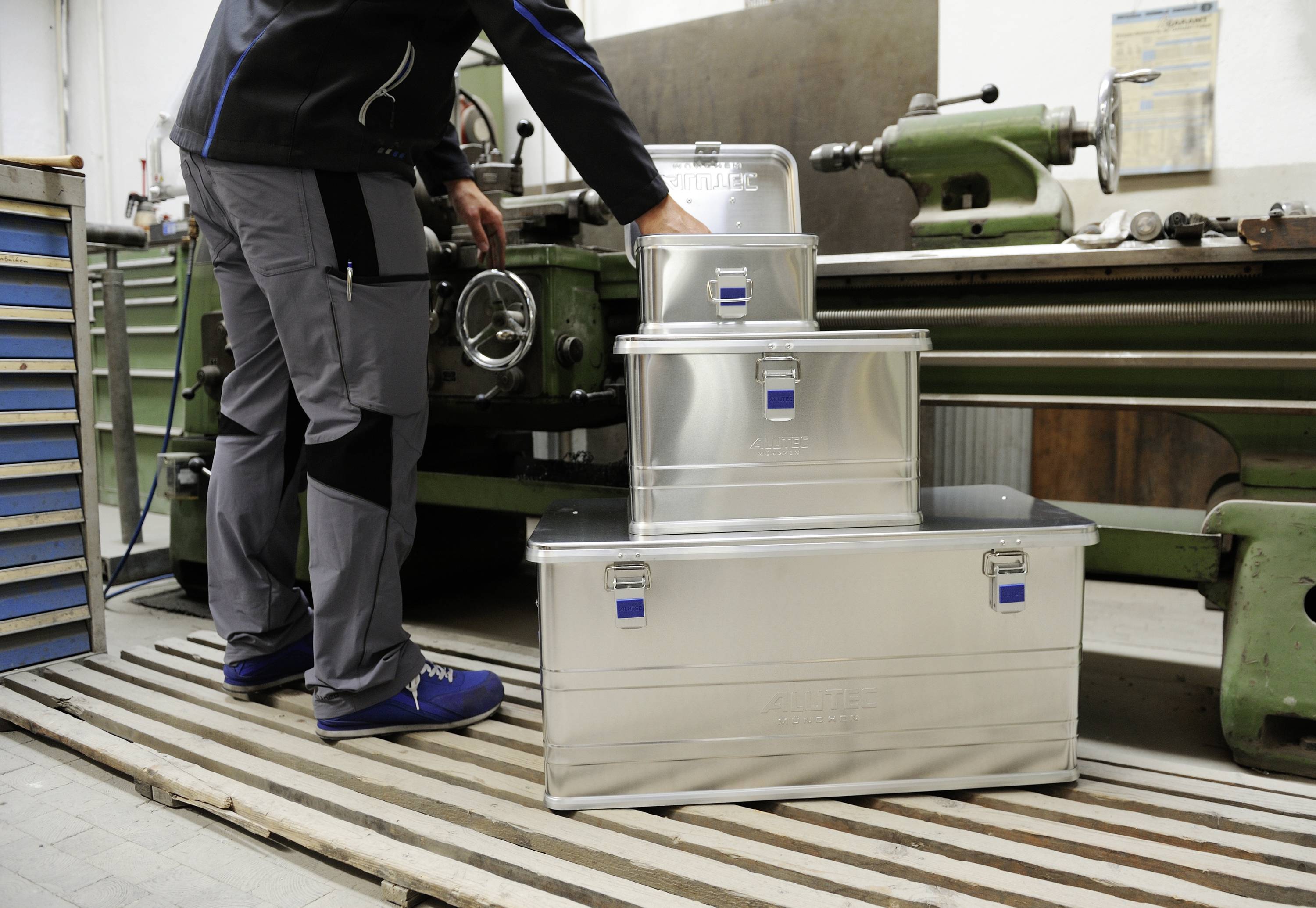 A person in workwear is placing a metal box on top of two other stacked boxes in front of an old, green machine tool in a workshop.