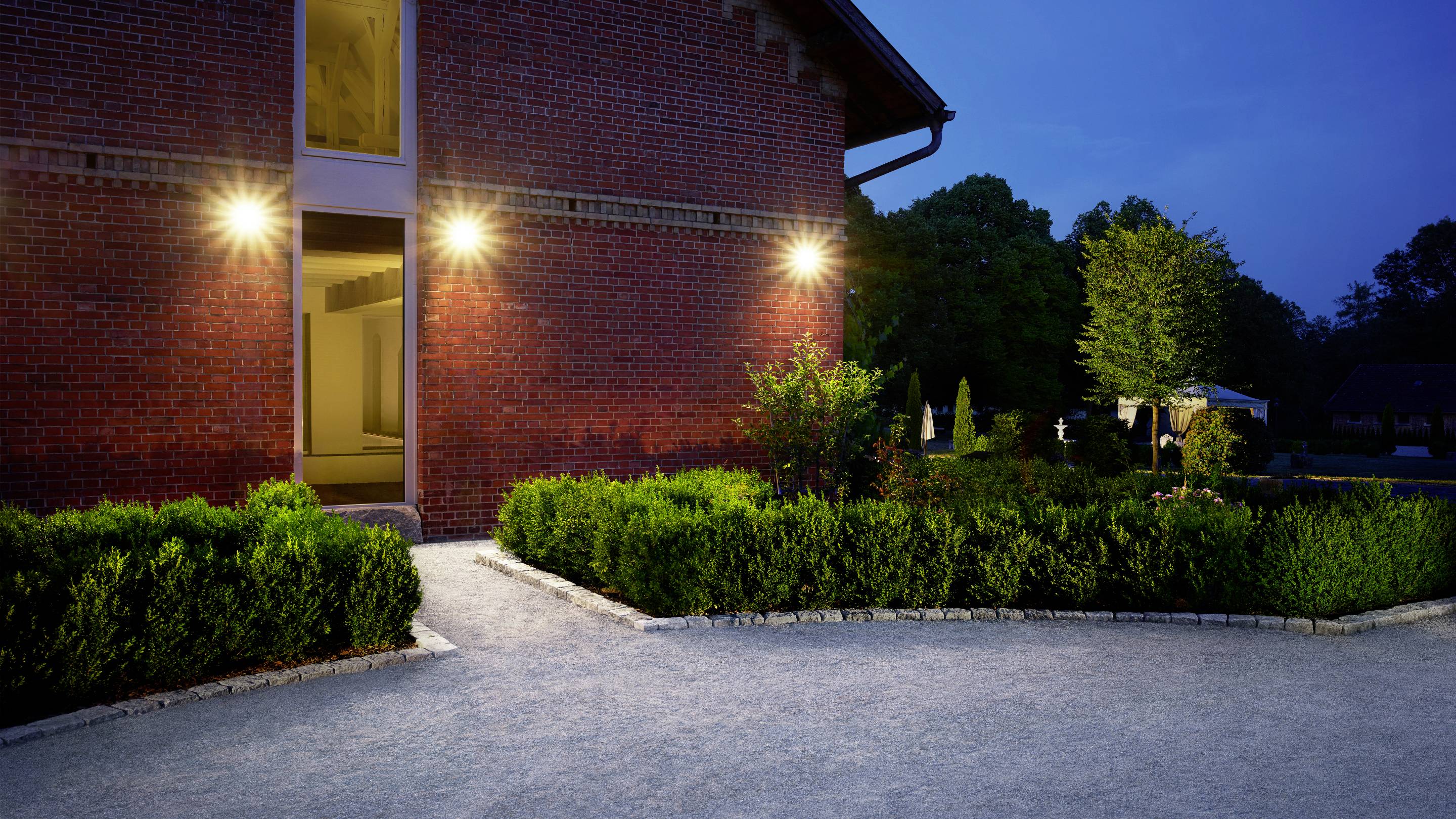 A illuminated entrance of a red brick building at dusk, surrounded by well-maintained hedges and a pathway, leads into the garden.