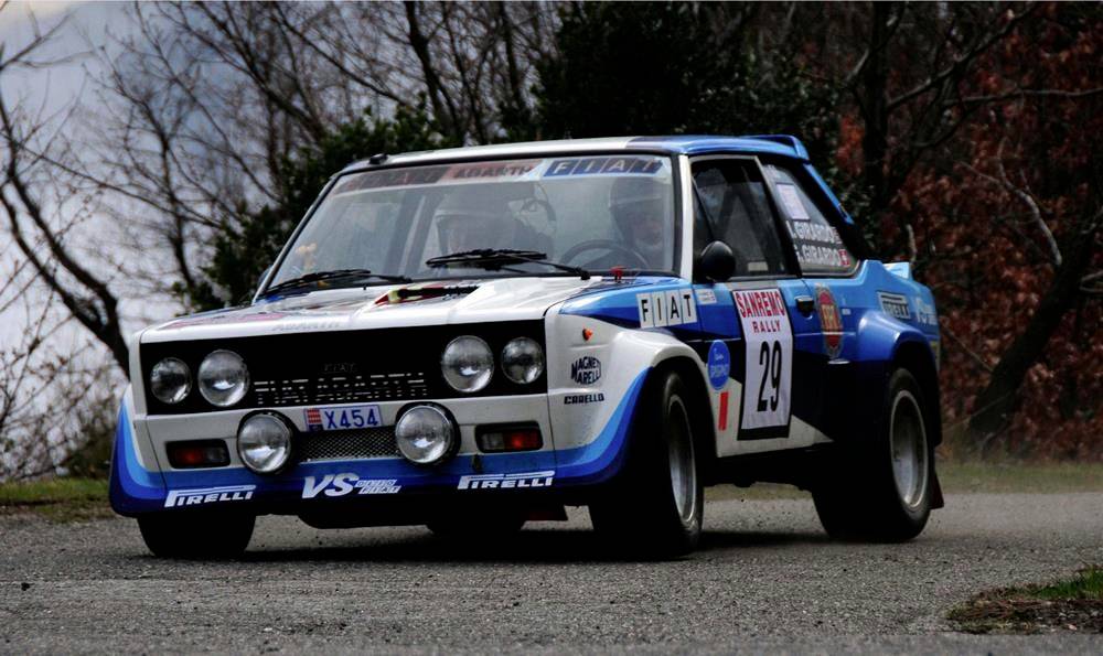 A rally car with the start number 29 speeds around a bend on a narrow, winding forest track.