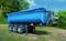 A blue trailer with three wheels stands on a meadow, surrounded by trees and shrubs. The sky is clear and blue.