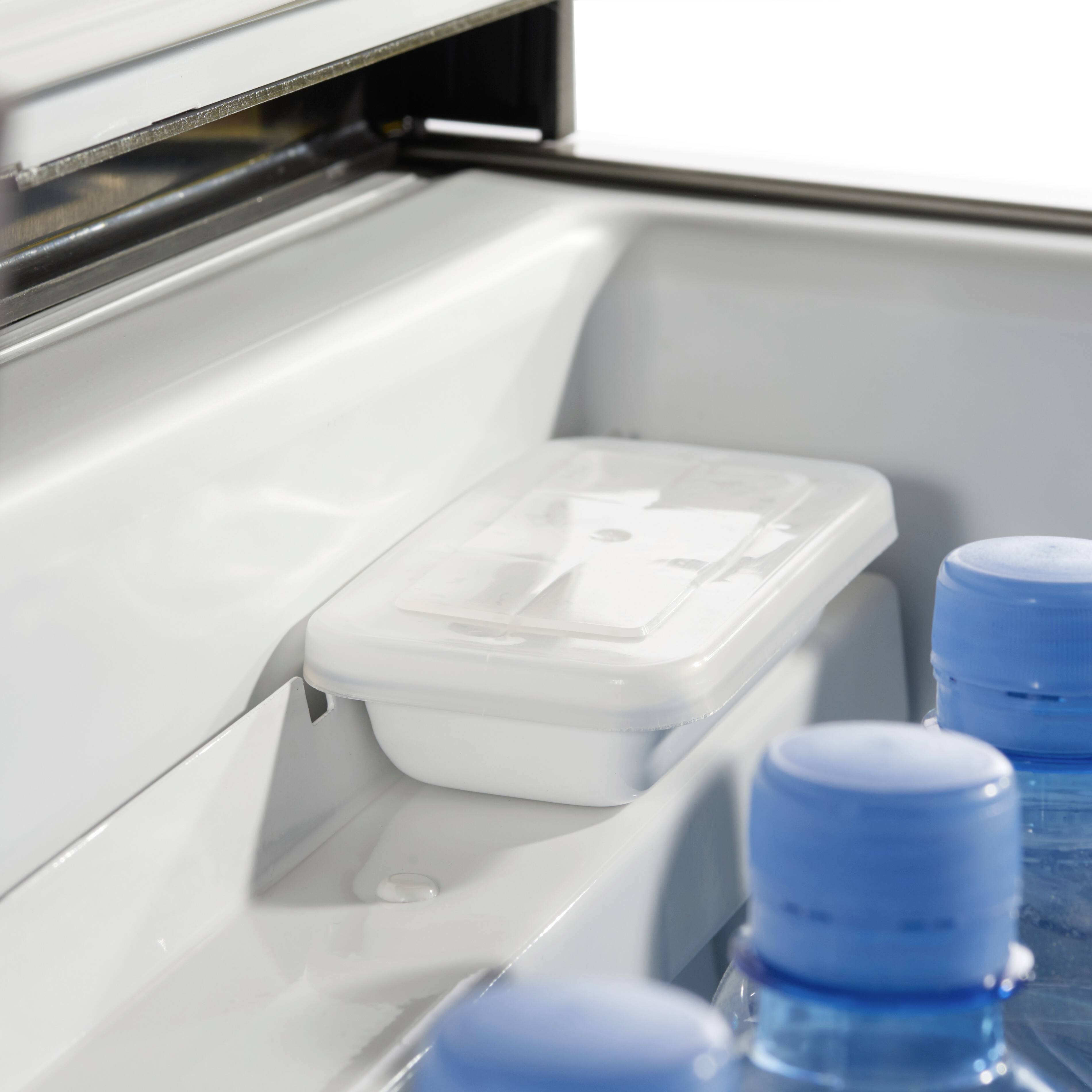 An open fridge reveals water bottles and a rectangular container sitting on a shelf. The contents of the container are not visible.