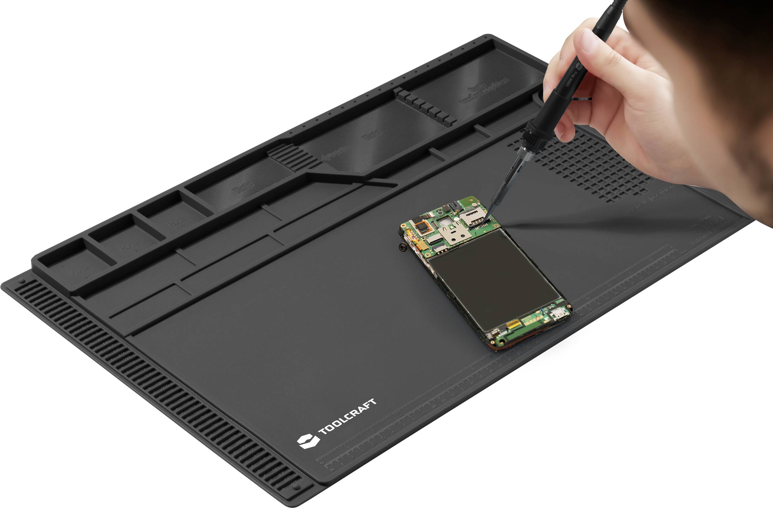 A person is repairing a small electronic circuit board with a soldering iron on a black, non-slip work mat with compartments.