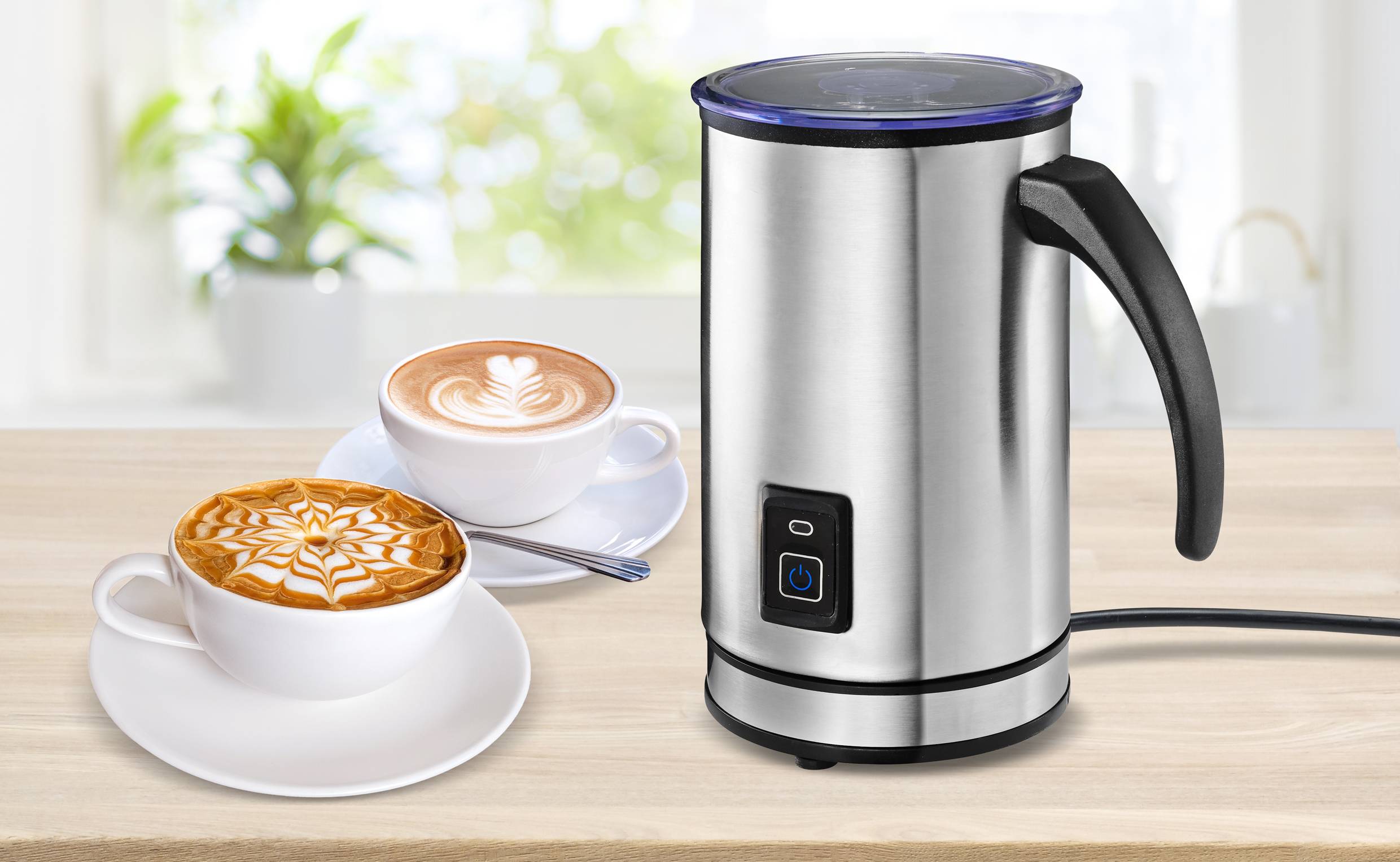 An electric milk frother sits on a kitchen worktop. Beside it are two cappuccinos with artistic milk foam patterns. In the background, a bright view through the window.