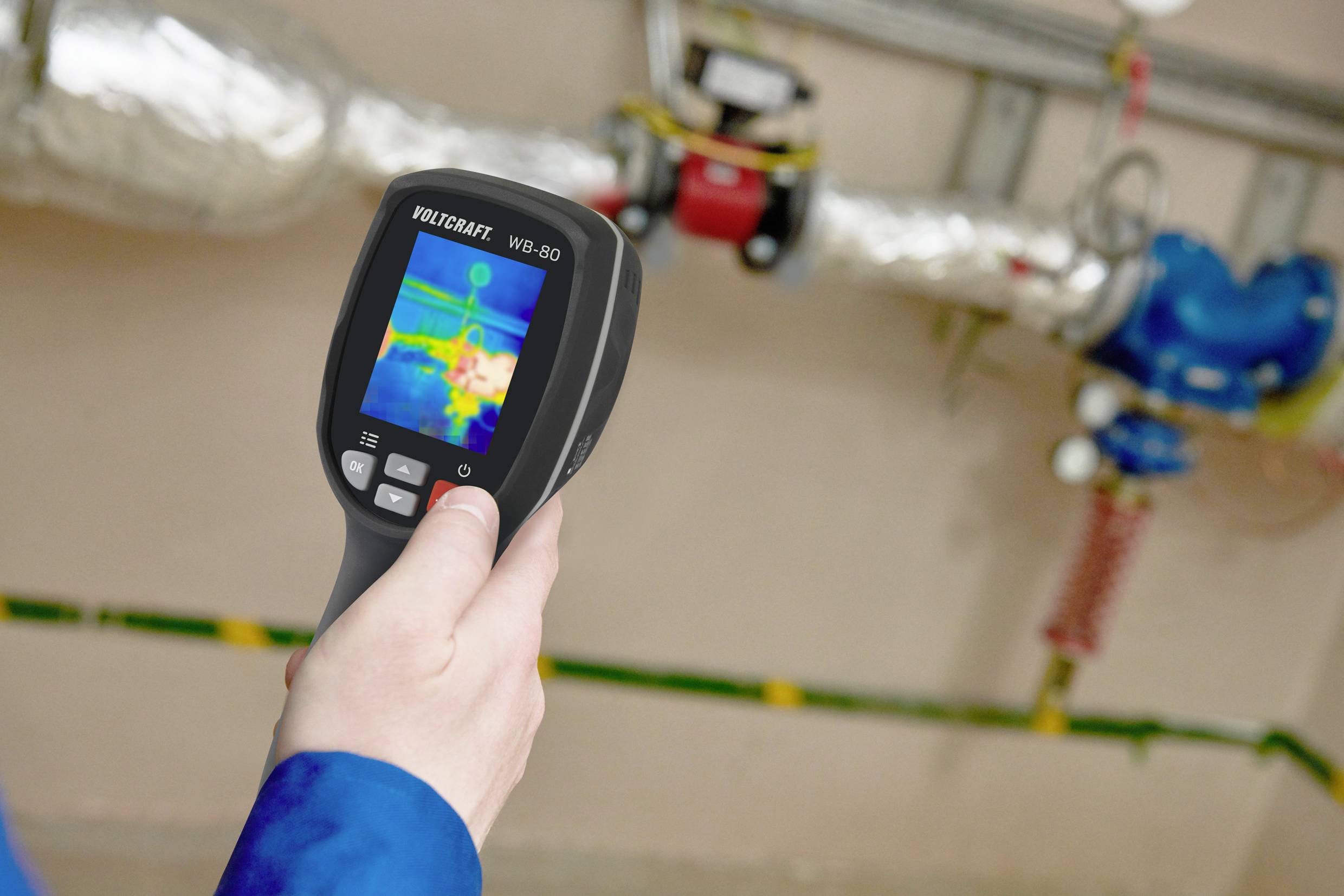 A person is holding a thermal imaging camera displaying a coloured thermal image of a heating pipe system to check the temperature distribution.