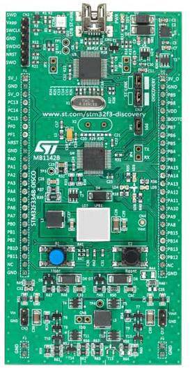 Microcontroller development board with a green printed circuit board, multiple electronic components, connectors, and labelling.