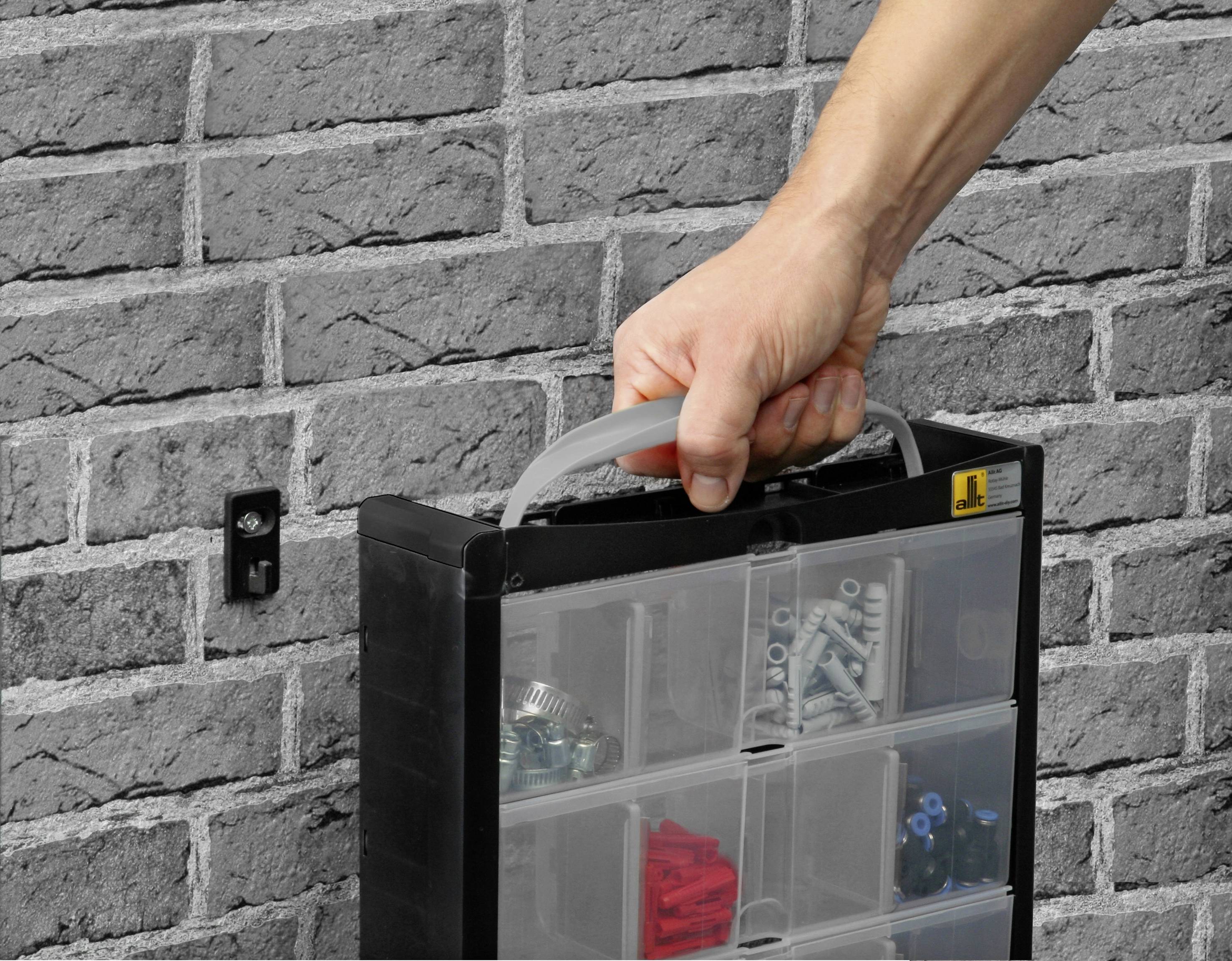A hand is holding a portable plastic organiser with compartments for screws and wall plugs. Background: grey brick wall.