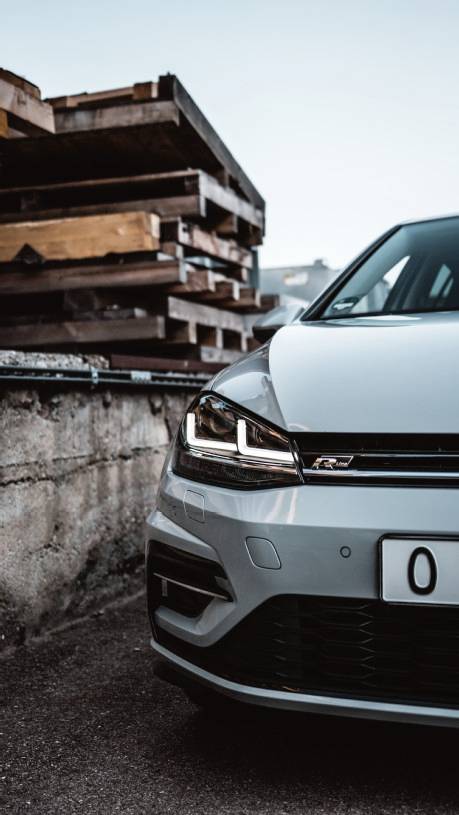 Front section of a silver car parked next to a stack of wooden pallets. The headlights are switched on, parked against a concrete wall.