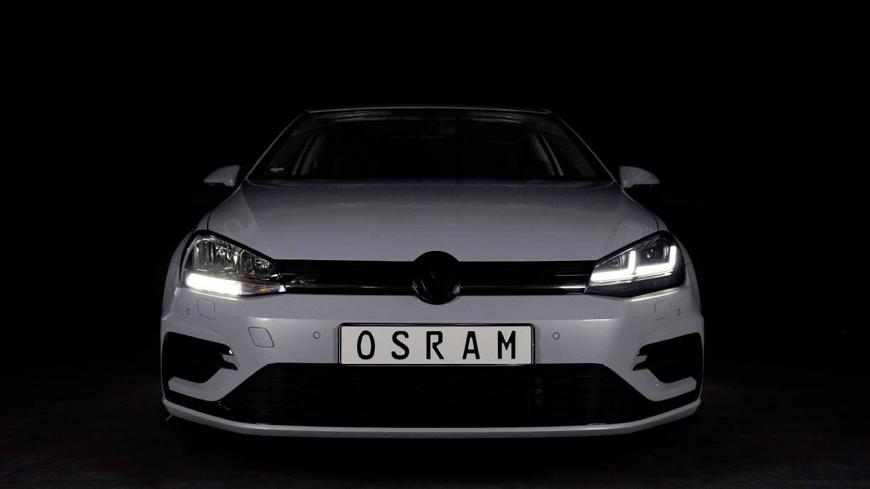 A white car with illuminated headlights is shown front-facing against a black background. The number plate reads 'OSRAM'.