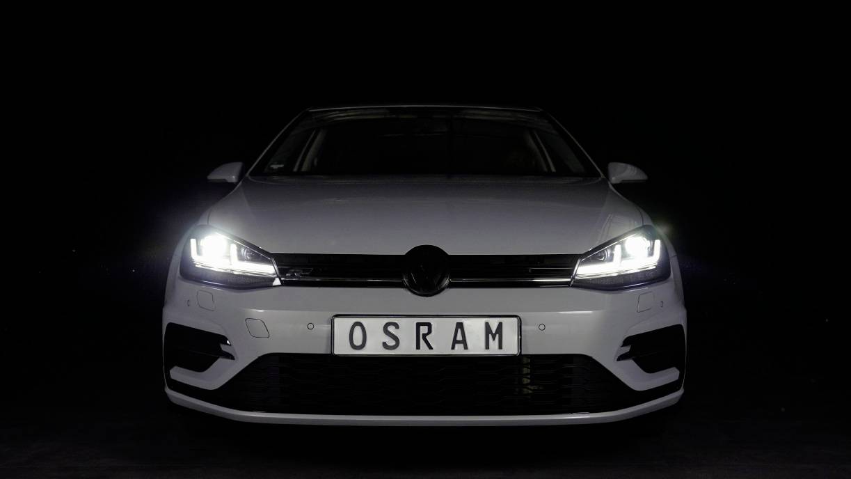 A white car, photographed head-on, against a black background. The number plate reads 'OSRAM'. The headlights are switched on.