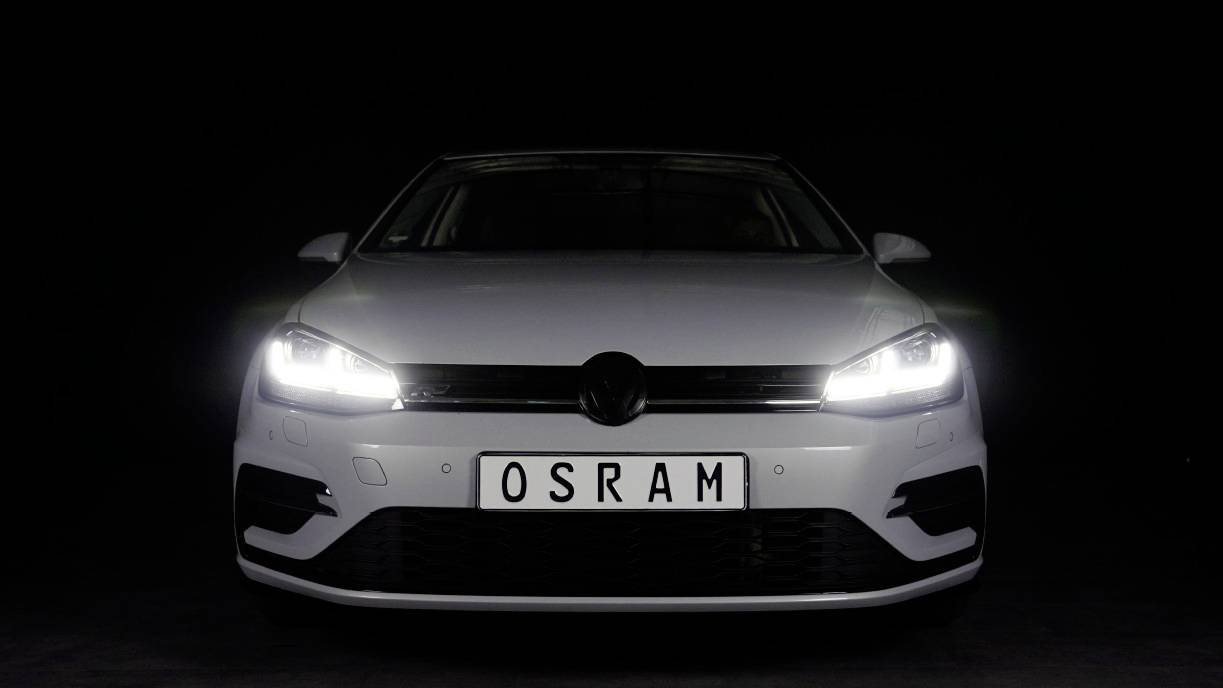 A white car with headlights switched on is positioned front-facing against a black background. The number plate reads 'OSRAM'.