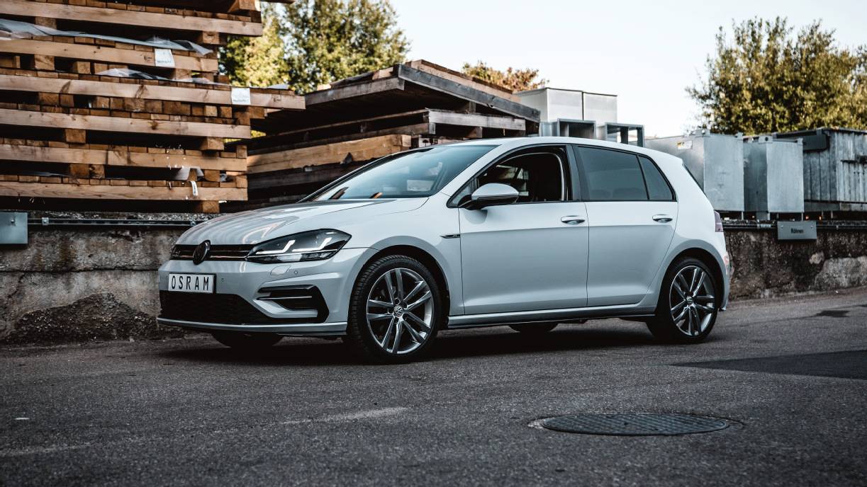 A silver compact car is parked on asphalt, surrounded by stacked timber and metal containers. The background shows trees and sky.