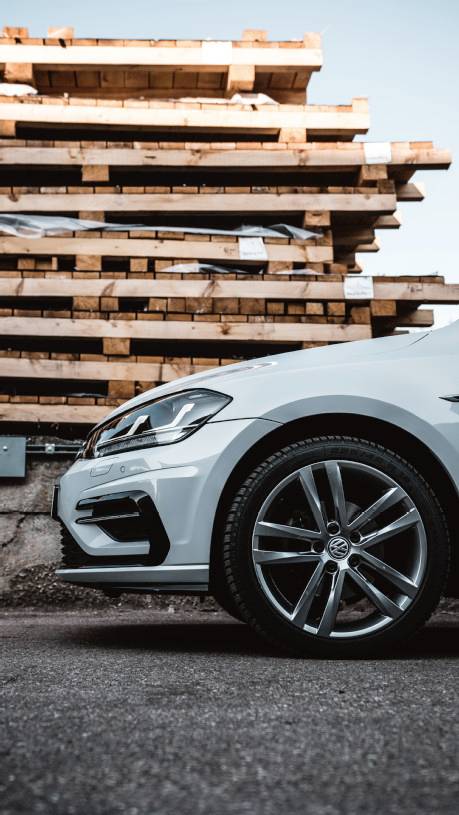 A white car is parked in front of a stack of wooden pallets. Only the front section of the car with one wheel is visible.