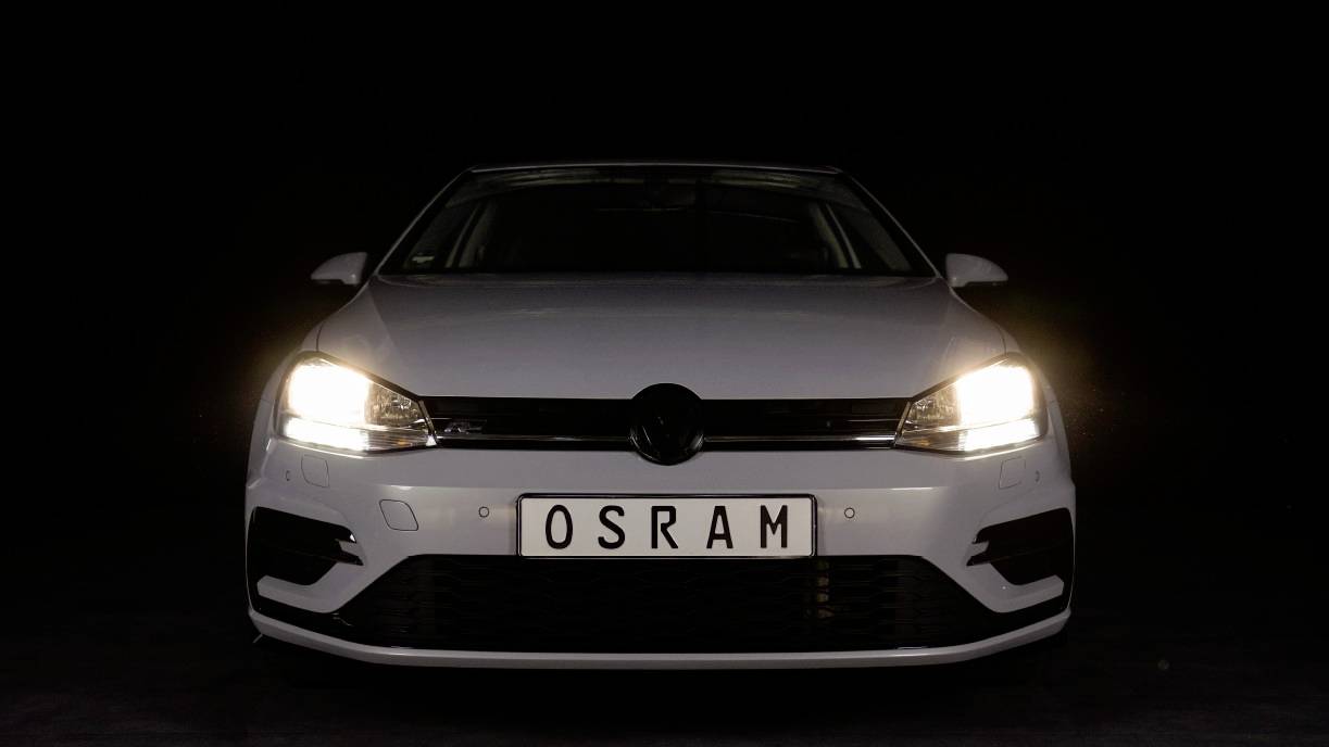 A silver car with headlights switched on in a dark environment, number plate reading 'OSRAM'.
