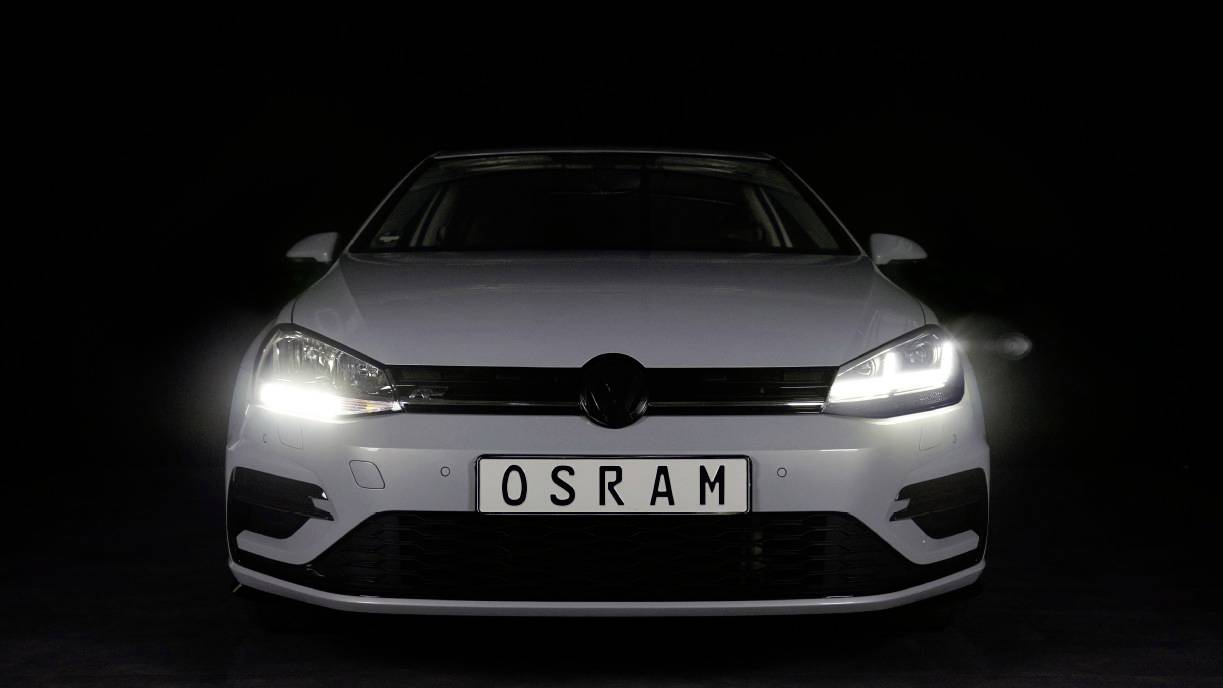 A white car with headlights switched on is shown head-on. The number plate reads 'OSRAM'.