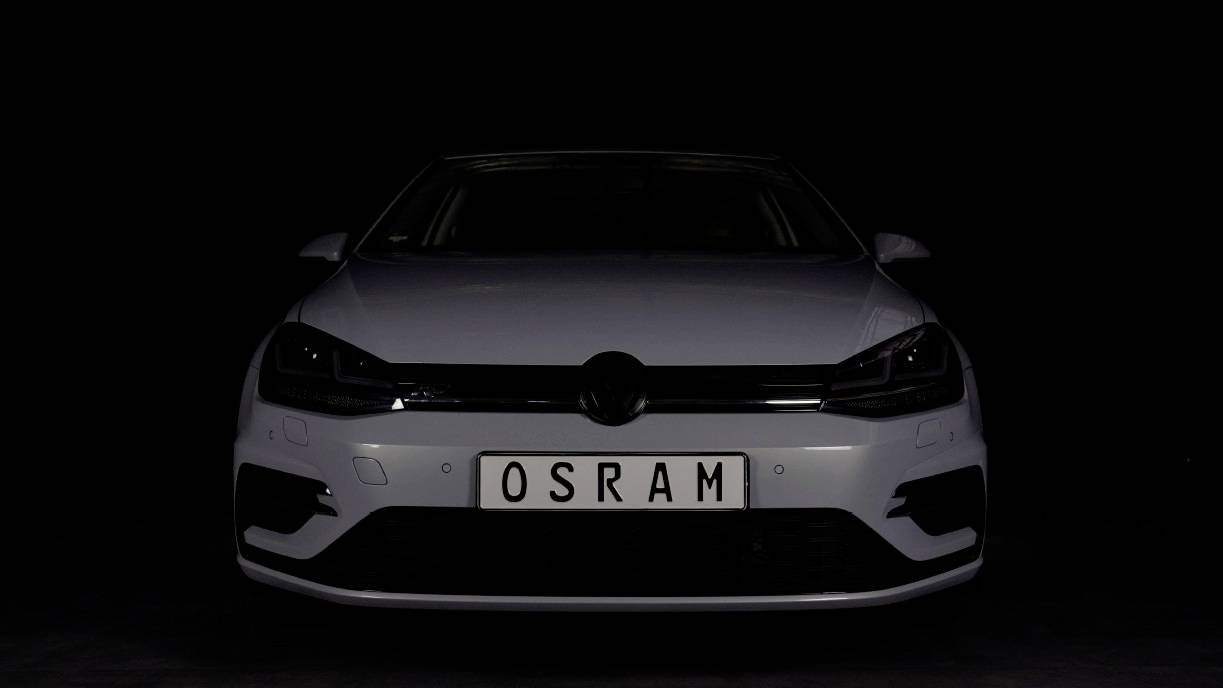 A white car in the darkness with a number plate 'OSRAM', viewed head-on. The focus is on the vehicle's lighting.