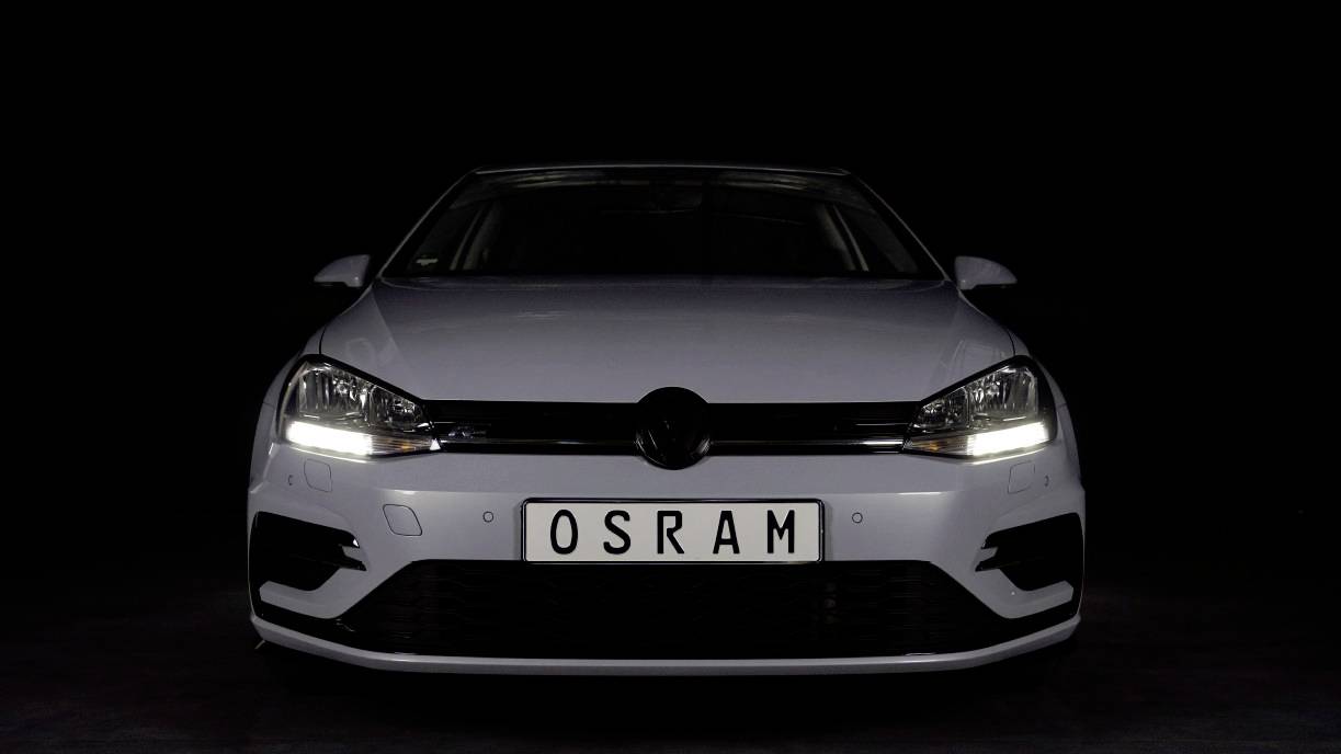 A white car with its headlight switched on is seen directly from the front. The number plate reads 'OSRAM'. Background is black.