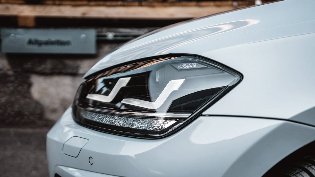 Close-up of a modern headlight of a white car, with a distinctive, stylised LED design, against a blurred background.
