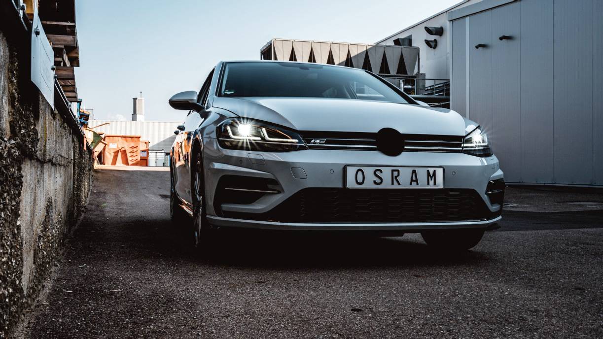 Car viewed from the front on a road, with a number plate reading 'OSRAM', illuminated headlights, industrial environment in the background.