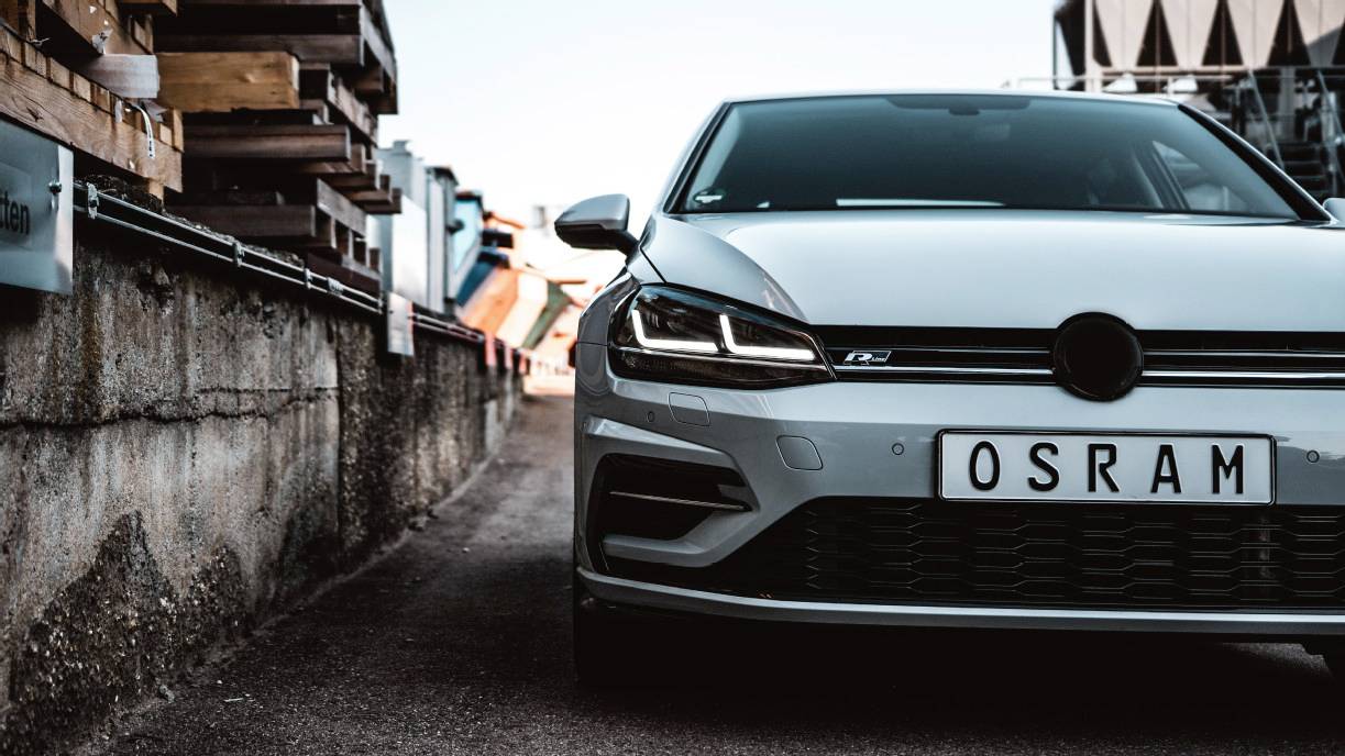 A silver car with the number plate 'OSRAM' is parked on an urban street beside a concrete wall in daylight.