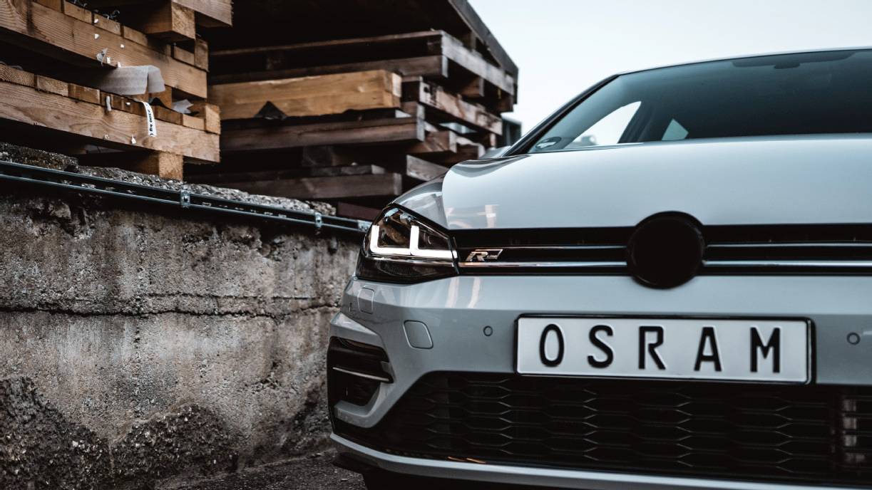 Car parked in front of a pile of wood and a wall, number plate reading 'OSRAM'. Front headlight design visible.