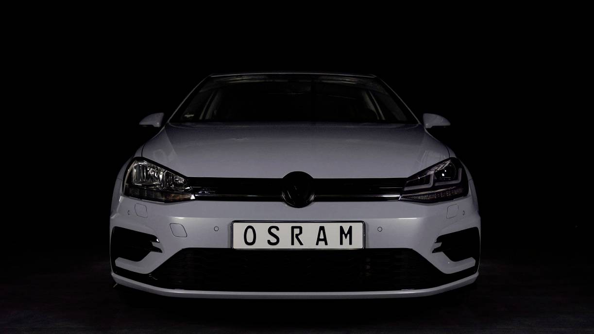A white car is parked in a dark room. The number plate reads 'OSRAM'. The car is depicted from a frontal view.