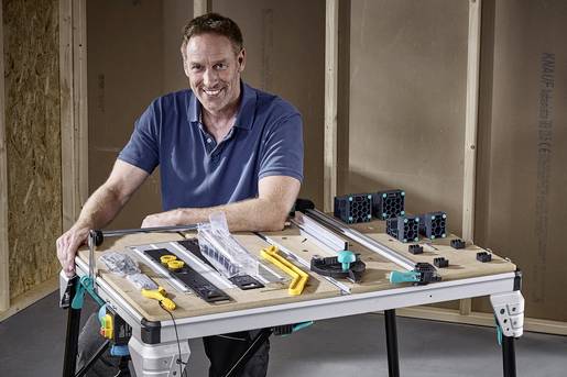 A man is standing smiling at a workbench with tools and accessories, in a room with partially completed walls.